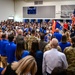 A large patriotic assembly is taking place inside a school gymnasium, with hundreds of students, parents, and military personnel gathered together. The scoreboard identifies the home team as the 'Aviators,' and flags including the American flag and the Arizona state flag are prominently displayed. Military personnel in camouflage uniforms and civilians in blue shirts are present, suggesting a Veterans Day or military appreciation event.