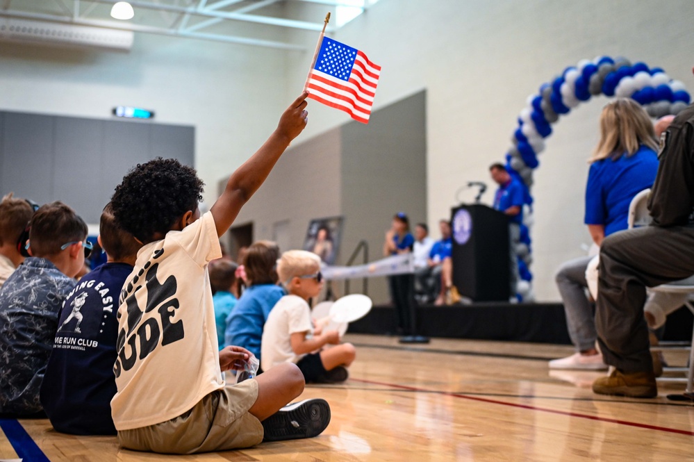 A young boy sits on a gymnasium floor among other children, enthusiastically raising a small American flag during what appears to be a school or community event. A stage with a podium, balloon arch in blue, white, and gray, and seated participants is visible in the background. The event appears to be a formal ceremony or assembly held indoors.