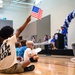 A young boy sits on a gymnasium floor among other children, enthusiastically raising a small American flag during what appears to be a school or community event. A stage with a podium, balloon arch in blue, white, and gray, and seated participants is visible in the background. The event appears to be a formal ceremony or assembly held indoors.