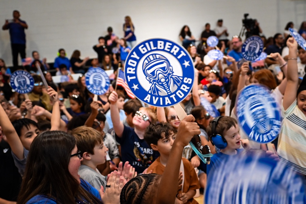 A large crowd of elementary school-aged children are gathered in what appears to be a school gymnasium or auditorium for a pep rally or school spirit event. The children are enthusiastically waving circular paddle signs bearing the 'Troy Gilbert Aviators' logo, which features a pilot mascot in blue and white. The event appears to be a school assembly celebrating the Troy Gilbert school mascot and team identity.