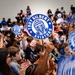 A large crowd of elementary school-aged children are gathered in what appears to be a school gymnasium or auditorium for a pep rally or school spirit event. The children are enthusiastically waving circular paddle signs bearing the 'Troy Gilbert Aviators' logo, which features a pilot mascot in blue and white. The event appears to be a school assembly celebrating the Troy Gilbert school mascot and team identity.