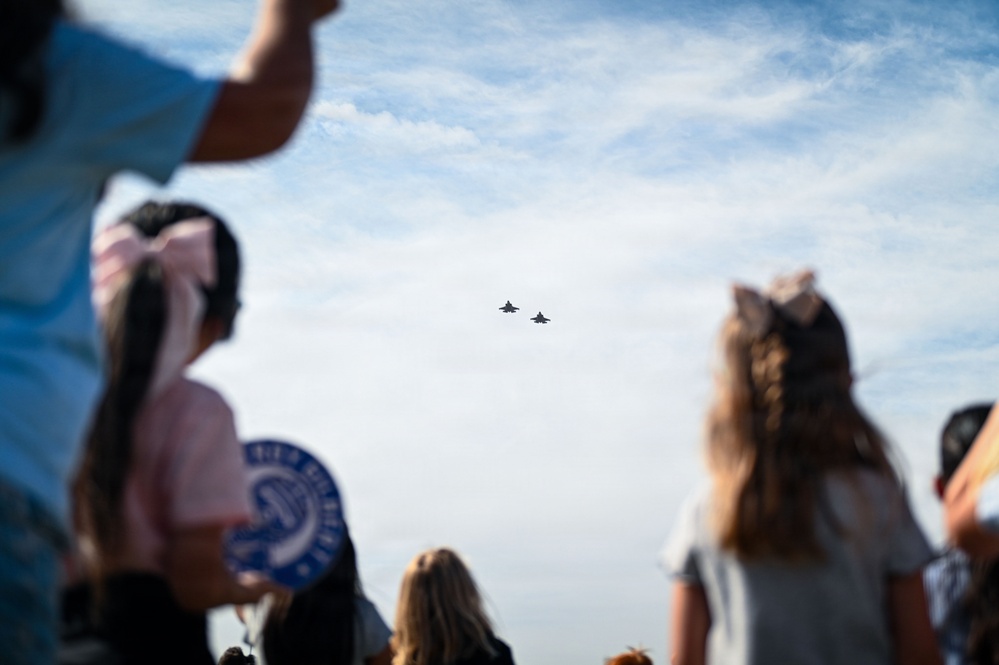 A crowd of spectators, including children and adults, watches two military jet aircraft flying in formation against a partly cloudy blue sky. The foreground figures are intentionally blurred using shallow depth of field, drawing focus to the aircraft in the background. The scene appears to be an outdoor air show or flyover event.