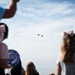 A crowd of spectators, including children and adults, watches two military jet aircraft flying in formation against a partly cloudy blue sky. The foreground figures are intentionally blurred using shallow depth of field, drawing focus to the aircraft in the background. The scene appears to be an outdoor air show or flyover event.