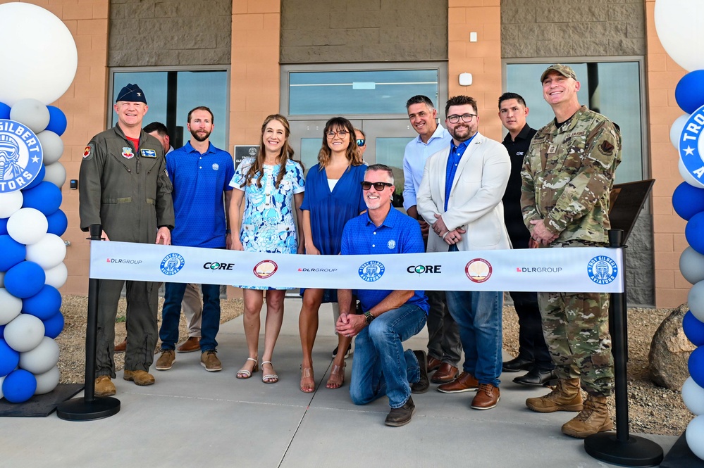 A ribbon-cutting ceremony is taking place in front of a building associated with the Troy Gilbert Aviators, featuring a group of approximately ten people including two military personnel in uniform. The ribbon displays logos for DLR Group, CORE, and Troy Gilbert Aviators. Blue and white balloon arches frame the entrance on both sides.