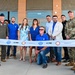 A ribbon-cutting ceremony is taking place in front of a building associated with the Troy Gilbert Aviators, featuring a group of approximately ten people including two military personnel in uniform. The ribbon displays logos for DLR Group, CORE, and Troy Gilbert Aviators. Blue and white balloon arches frame the entrance on both sides.