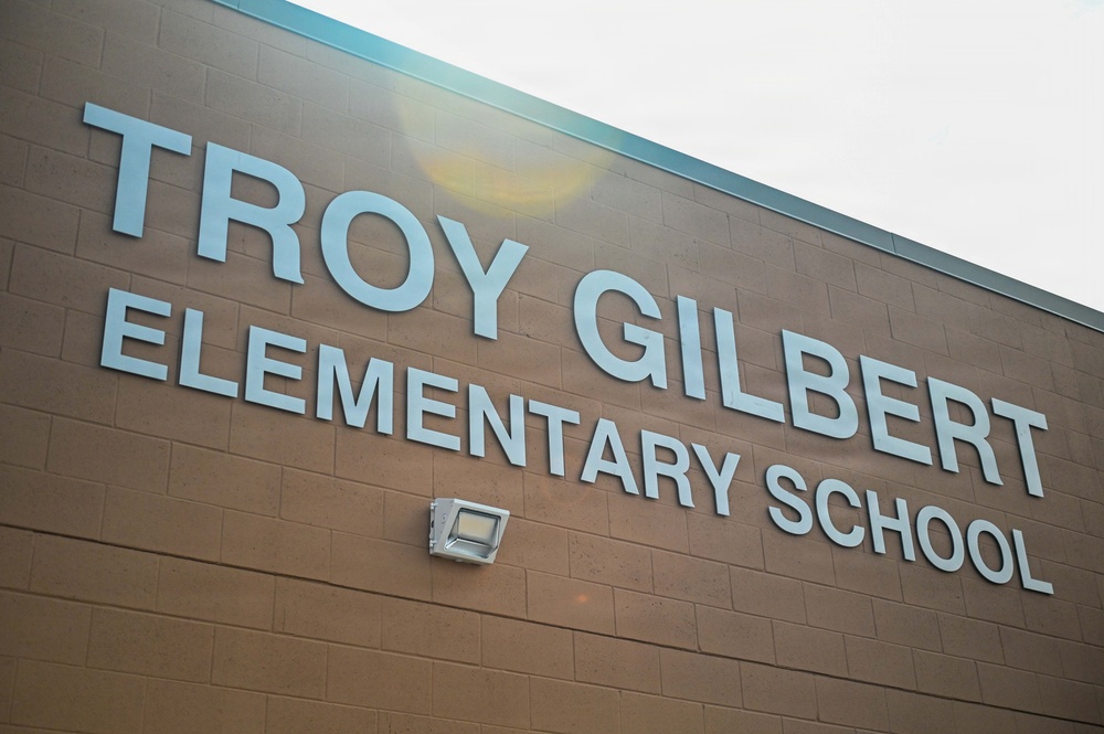 The image shows the exterior facade of Troy Gilbert Elementary School, featuring large light-blue dimensional letters mounted on a brown brick wall. A wall-mounted flood light fixture is visible below the signage. The photo is taken from a low angle looking upward, with an overcast sky visible in the upper portion of the frame.