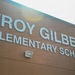 The image shows the exterior facade of Troy Gilbert Elementary School, featuring large light-blue dimensional letters mounted on a brown brick wall. A wall-mounted flood light fixture is visible below the signage. The photo is taken from a low angle looking upward, with an overcast sky visible in the upper portion of the frame.
