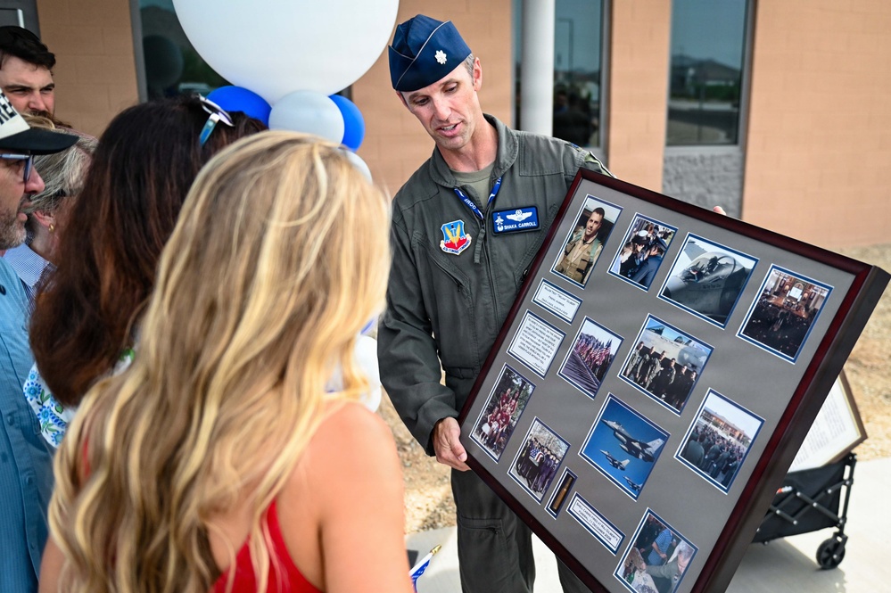 A U.S. Air Force officer in a flight suit and service cap is presenting a framed photo collage to a woman with blonde hair at what appears to be a military ceremony or retirement event. The framed display contains multiple photographs documenting what appears to be a military career, including images of aircraft and group photos. Blue and white balloons are visible in the background, suggesting a celebratory occasion.