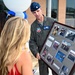 A U.S. Air Force officer in a flight suit and service cap is presenting a framed photo collage to a woman with blonde hair at what appears to be a military ceremony or retirement event. The framed display contains multiple photographs documenting what appears to be a military career, including images of aircraft and group photos. Blue and white balloons are visible in the background, suggesting a celebratory occasion.