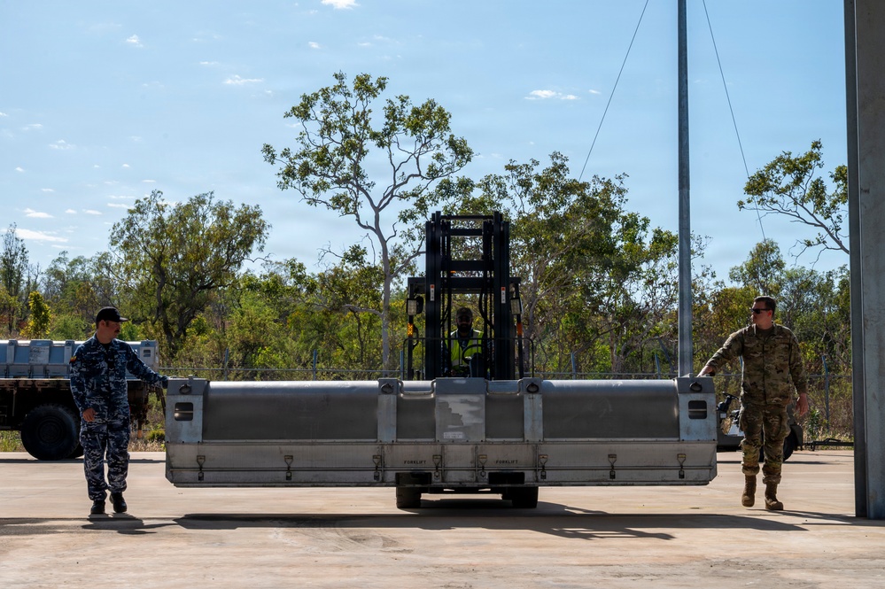 A forklift operator is maneuvering a large metal container or equipment pallet on a concrete apron, likely at a military airfield or logistics facility. Two military personnel — one in blue camouflage uniform and one in green/tan camouflage — are walking alongside the load, appearing to guide or spot the operation. The setting features sparse Australian-style eucalyptus vegetation in the background, suggesting a tropical or semi-arid military installation.