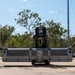 A forklift operator is maneuvering a large metal container or equipment pallet on a concrete apron, likely at a military airfield or logistics facility. Two military personnel — one in blue camouflage uniform and one in green/tan camouflage — are walking alongside the load, appearing to guide or spot the operation. The setting features sparse Australian-style eucalyptus vegetation in the background, suggesting a tropical or semi-arid military installation.