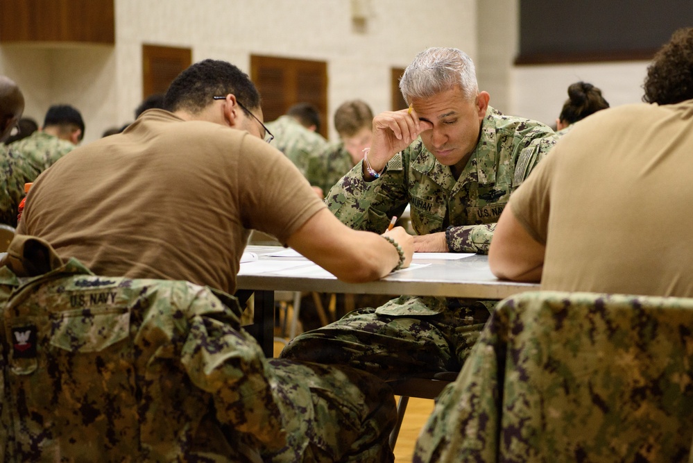 A group of U.S. Navy personnel are seated at tables in what appears to be a large hall or gymnasium, engaged in a written exercise or examination. The central figure is a senior Navy officer in NWU (Navy Working Uniform) camouflage, appearing focused and contemplative while writing. Multiple sailors in various uniform configurations are visible throughout the room.