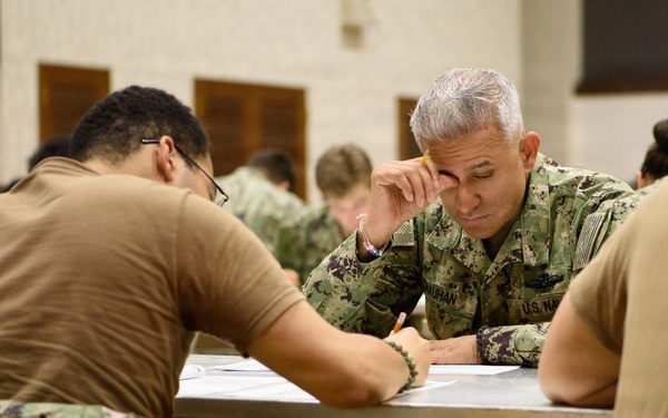 A group of U.S. Navy personnel are seated at tables in what appears to be a large hall or gymnasium, engaged in a written exercise or examination. The central figure is a senior Navy officer in NWU (Navy Working Uniform) camouflage, appearing focused and contemplative while writing. Multiple sailors in various uniform configurations are visible throughout the room.