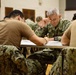 A group of U.S. Navy personnel are seated at tables in what appears to be a large hall or gymnasium, engaged in a written exercise or examination. The central figure is a senior Navy officer in NWU (Navy Working Uniform) camouflage, appearing focused and contemplative while writing. Multiple sailors in various uniform configurations are visible throughout the room.