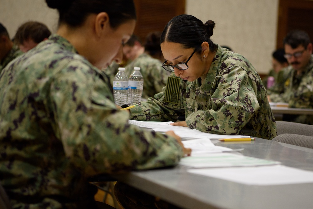 A group of military personnel in U.S. Navy Working Uniform (NWU Type III) camouflage are seated at tables completing written paperwork or an examination. The setting appears to be an indoor classroom or briefing room. Multiple service members are focused on their documents, with water bottles and pencils visible on the tables.