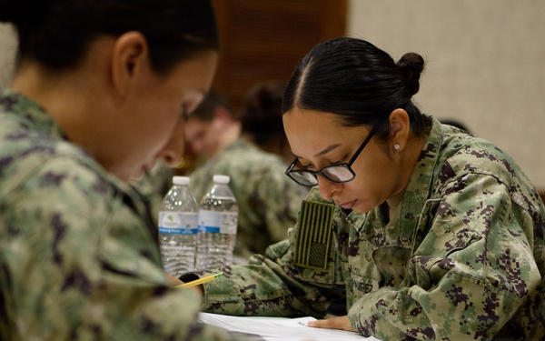 A group of military personnel in U.S. Navy Working Uniform (NWU Type III) camouflage are seated at tables completing written paperwork or an examination. The setting appears to be an indoor classroom or briefing room. Multiple service members are focused on their documents, with water bottles and pencils visible on the tables.