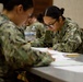 A group of military personnel in U.S. Navy Working Uniform (NWU Type III) camouflage are seated at tables completing written paperwork or an examination. The setting appears to be an indoor classroom or briefing room. Multiple service members are focused on their documents, with water bottles and pencils visible on the tables.