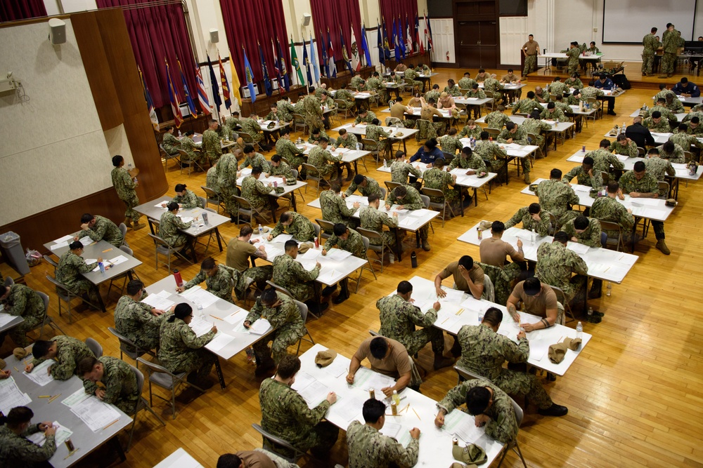 A large group of military personnel in camouflage uniforms are seated at individual desks in a spacious hall, appearing to take a written examination or complete official paperwork. The setting resembles a formal testing environment with rows of tables arranged across a polished wooden floor. Multiple flags are displayed along the back wall, and a few supervisory personnel are standing and observing.