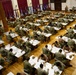 A large group of military personnel in camouflage uniforms are seated at individual desks in a spacious hall, appearing to take a written examination or complete official paperwork. The setting resembles a formal testing environment with rows of tables arranged across a polished wooden floor. Multiple flags are displayed along the back wall, and a few supervisory personnel are standing and observing.