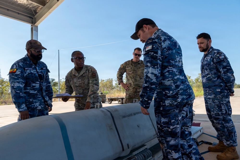 Military personnel from what appears to be the United States Air Force and the Royal Australian Air Force are gathered around a large munition or ordnance item on a tarmac or airfield surface. The group appears to be conducting an inspection, briefing, or joint training exercise involving the weapon system. The setting is an outdoor covered area with clear skies and vegetation visible in the background.