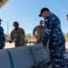Military personnel from what appears to be the United States Air Force and the Royal Australian Air Force are gathered around a large munition or ordnance item on a tarmac or airfield surface. The group appears to be conducting an inspection, briefing, or joint training exercise involving the weapon system. The setting is an outdoor covered area with clear skies and vegetation visible in the background.