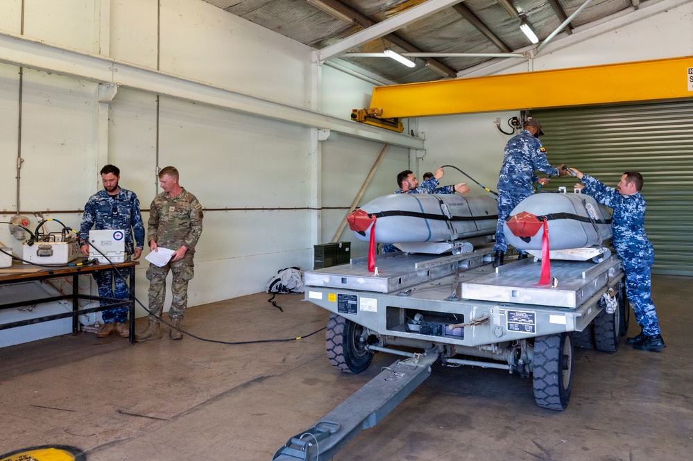 Military personnel are working inside a hangar or maintenance facility, handling what appear to be aerial munitions or ordnance items loaded on a wheeled cart or trailer. The group includes individuals in blue digital camouflage uniforms and one person in a multicam/OCP pattern uniform. The setting suggests a weapons loading, inspection, or maintenance operation.