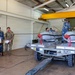 Military personnel are working inside a hangar or maintenance facility, handling what appear to be aerial munitions or ordnance items loaded on a wheeled cart or trailer. The group includes individuals in blue digital camouflage uniforms and one person in a multicam/OCP pattern uniform. The setting suggests a weapons loading, inspection, or maintenance operation.
