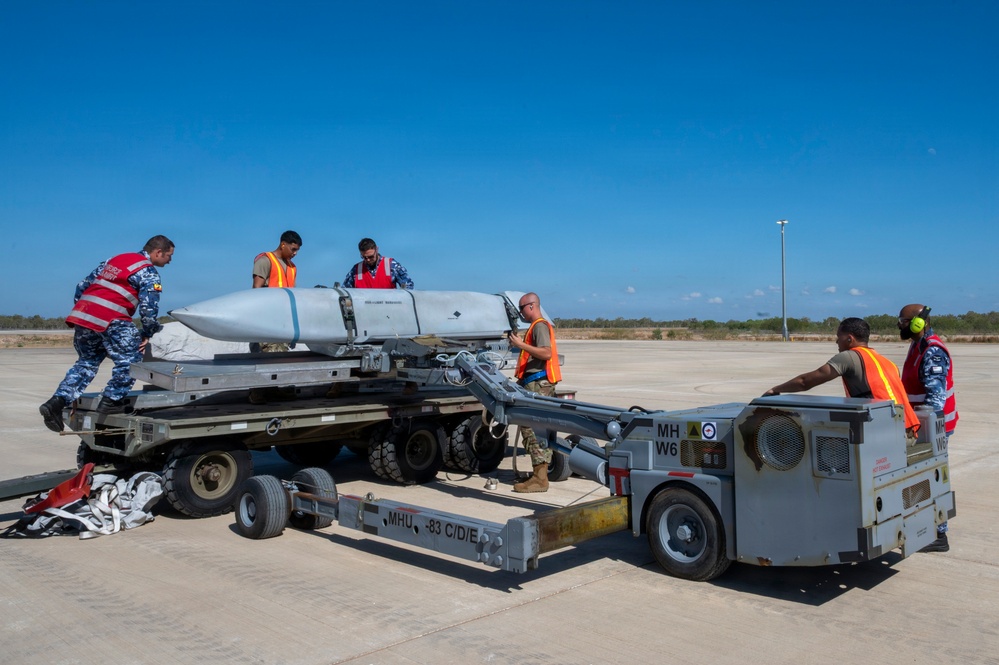 Military personnel are handling a large missile or munition on a weapons loading cart (MHU-83 C/D/E) on an airfield tarmac. The team consists of approximately six individuals wearing high-visibility orange vests and red safety vests, working together to manage the ordnance. The equipment and markings suggest a joint or multinational military exercise or routine weapons handling operation.