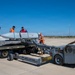 Military personnel are handling a large missile or munition on a weapons loading cart (MHU-83 C/D/E) on an airfield tarmac. The team consists of approximately six individuals wearing high-visibility orange vests and red safety vests, working together to manage the ordnance. The equipment and markings suggest a joint or multinational military exercise or routine weapons handling operation.
