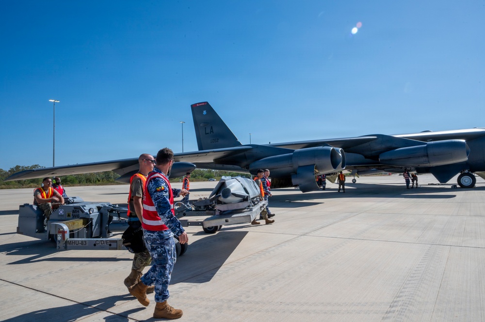 Military personnel in safety vests are conducting weapons loading operations on a B-52 Stratofortress bomber on an airfield tarmac. The aircraft displays 'LA' tail codes indicating it belongs to the 2nd Bomb Wing based at Barksdale Air Force Base, Louisiana. Ground crew members are maneuvering what appears to be a munition or weapons system on an MHU-83 munitions handling unit toward the aircraft.