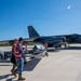Military personnel in safety vests are conducting weapons loading operations on a B-52 Stratofortress bomber on an airfield tarmac. The aircraft displays 'LA' tail codes indicating it belongs to the 2nd Bomb Wing based at Barksdale Air Force Base, Louisiana. Ground crew members are maneuvering what appears to be a munition or weapons system on an MHU-83 munitions handling unit toward the aircraft.