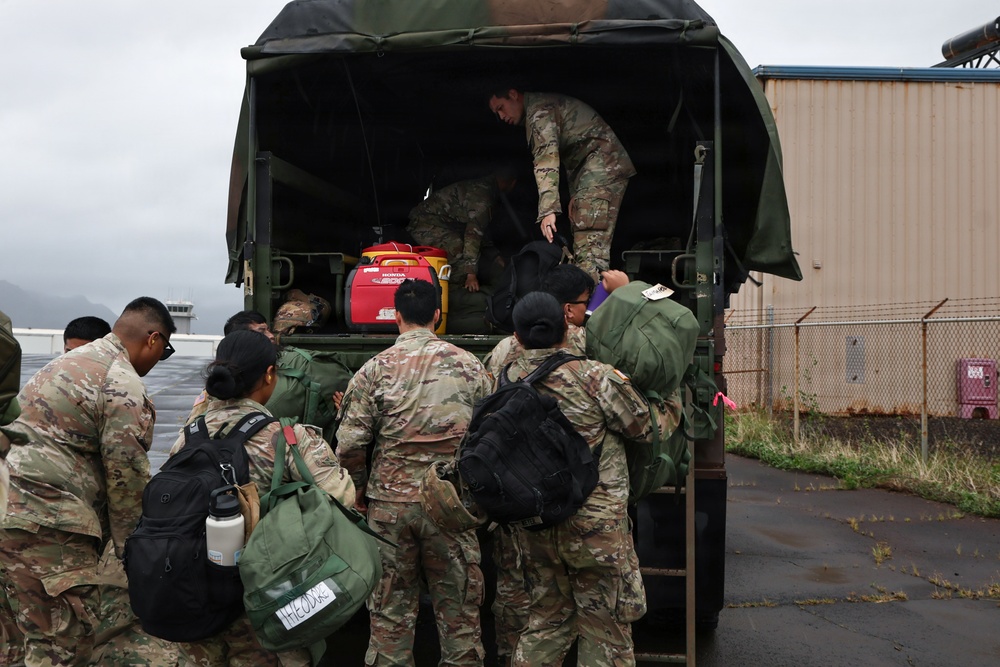 A group of military personnel in OCP (Operational Camouflage Pattern) uniforms are loading or unloading equipment and gear from the back of a military canvas-covered truck. One soldier is positioned inside the truck bed assisting with the transfer of items, while others stand at the rear handing up or receiving bags and equipment. The scene appears to take place on a wet tarmac near a military or airfield facility under overcast skies.