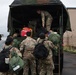 A group of military personnel in OCP (Operational Camouflage Pattern) uniforms are loading or unloading equipment and gear from the back of a military canvas-covered truck. One soldier is positioned inside the truck bed assisting with the transfer of items, while others stand at the rear handing up or receiving bags and equipment. The scene appears to take place on a wet tarmac near a military or airfield facility under overcast skies.