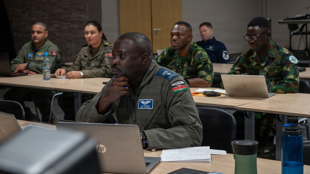 A multinational military classroom setting showing personnel from multiple nations attending what appears to be a joint training or educational seminar. The foreground features a Kenya Air Force officer in a flight suit with visible name tape and national flag patch. Several other military personnel in various national uniforms are seated at desks with laptops in the background.