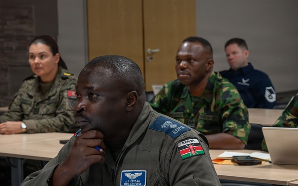 A multinational military classroom setting showing personnel from multiple nations attending what appears to be a joint training or educational seminar. The foreground features a Kenya Air Force officer in a flight suit with visible name tape and national flag patch. Several other military personnel in various national uniforms are seated at desks with laptops in the background.