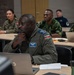 A multinational military classroom setting showing personnel from multiple nations attending what appears to be a joint training or educational seminar. The foreground features a Kenya Air Force officer in a flight suit with visible name tape and national flag patch. Several other military personnel in various national uniforms are seated at desks with laptops in the background.