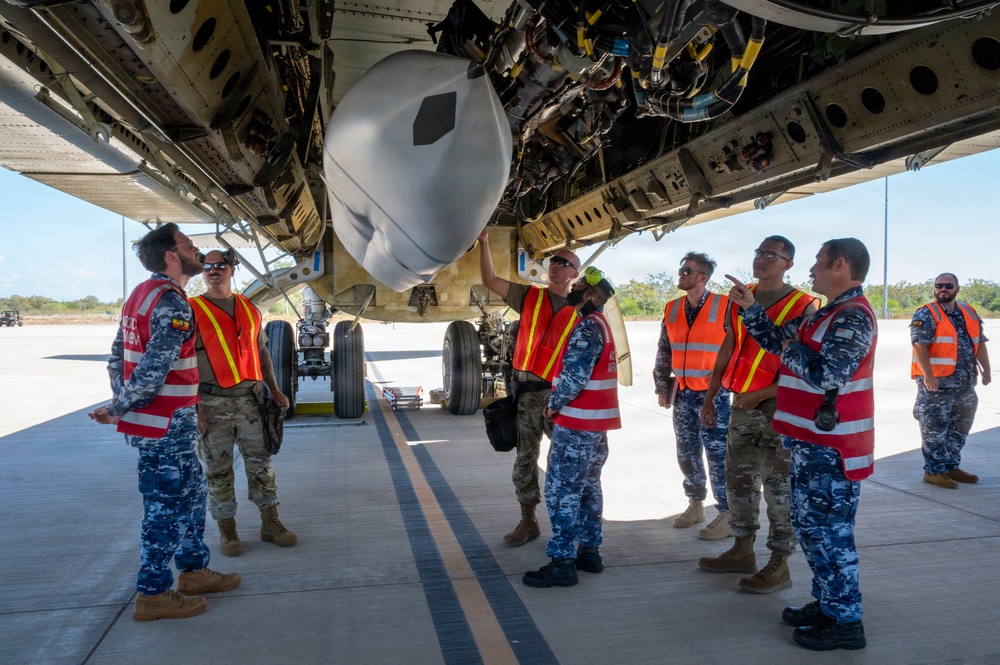 A group of military personnel wearing orange high-visibility safety vests are gathered beneath a large military aircraft on a tarmac, conducting what appears to be an inspection or briefing. The group includes individuals in both camouflage uniforms and blue digital-pattern uniforms, suggesting a multinational or joint-service exercise. One person is pointing at or touching a component on the underside of the aircraft while others observe.