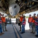 A group of military personnel wearing orange high-visibility safety vests are gathered beneath a large military aircraft on a tarmac, conducting what appears to be an inspection or briefing. The group includes individuals in both camouflage uniforms and blue digital-pattern uniforms, suggesting a multinational or joint-service exercise. One person is pointing at or touching a component on the underside of the aircraft while others observe.