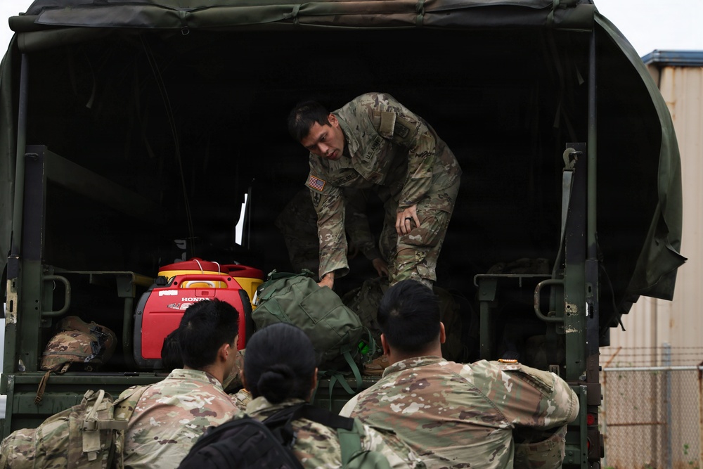 A group of U.S. military personnel in OCP (Operational Camouflage Pattern) uniforms are loading or unloading equipment from the rear of a military tactical vehicle with a green canvas cover. One soldier is positioned inside the truck bed, handling a large duffel bag, while several others stand at the rear assisting. A red Honda generator and other military gear are visible inside the truck.