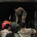A group of U.S. military personnel in OCP (Operational Camouflage Pattern) uniforms are loading or unloading equipment from the rear of a military tactical vehicle with a green canvas cover. One soldier is positioned inside the truck bed, handling a large duffel bag, while several others stand at the rear assisting. A red Honda generator and other military gear are visible inside the truck.