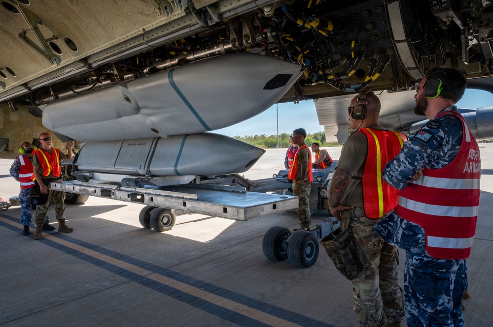 Military armament personnel are loading or inspecting large munitions beneath the fuselage of a heavy bomber aircraft on a flight line. The team includes both US Air Force personnel in multicam uniforms and what appear to be Royal Australian Air Force (RAAF) armament technicians wearing distinctive blue camouflage uniforms. All personnel are wearing orange high-visibility safety vests, indicating an active ordnance handling operation.