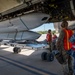 Military armament personnel are loading or inspecting large munitions beneath the fuselage of a heavy bomber aircraft on a flight line. The team includes both US Air Force personnel in multicam uniforms and what appear to be Royal Australian Air Force (RAAF) armament technicians wearing distinctive blue camouflage uniforms. All personnel are wearing orange high-visibility safety vests, indicating an active ordnance handling operation.