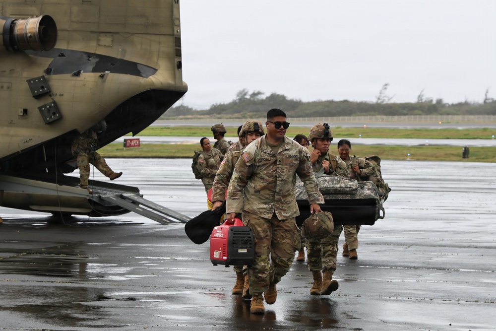 A group of U.S. Army soldiers in OCP (Operational Camouflage Pattern) uniforms are unloading equipment from what appears to be a CH-47 Chinook helicopter on a wet tarmac during overcast, windy conditions. The lead soldier is carrying a red portable generator and additional gear, while others behind carry various equipment including what appears to be a folded cot or stretcher. The scene suggests a disaster relief or emergency response deployment.