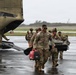 A group of U.S. Army soldiers in OCP (Operational Camouflage Pattern) uniforms are unloading equipment from what appears to be a CH-47 Chinook helicopter on a wet tarmac during overcast, windy conditions. The lead soldier is carrying a red portable generator and additional gear, while others behind carry various equipment including what appears to be a folded cot or stretcher. The scene suggests a disaster relief or emergency response deployment.