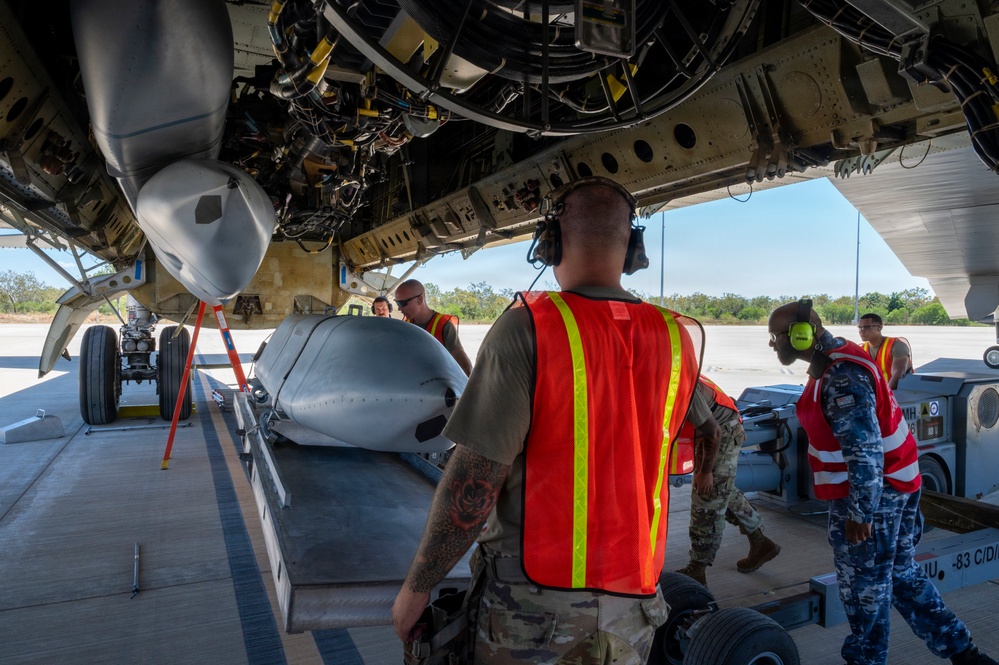 Military personnel are conducting weapons or munitions loading operations beneath a large aircraft, likely a bomber. Ground crew members wearing high-visibility orange safety vests and hearing protection are working alongside what appears to be a large munition or ordnance on a loading cart positioned under the aircraft's open bomb bay. Personnel in both U.S. military camouflage uniforms and what appears to be a foreign military uniform are present, suggesting a joint or coalition operation.