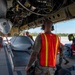 Military personnel are conducting weapons or munitions loading operations beneath a large aircraft, likely a bomber. Ground crew members wearing high-visibility orange safety vests and hearing protection are working alongside what appears to be a large munition or ordnance on a loading cart positioned under the aircraft's open bomb bay. Personnel in both U.S. military camouflage uniforms and what appears to be a foreign military uniform are present, suggesting a joint or coalition operation.