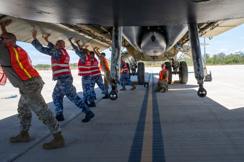 Military personnel from what appears to be a joint multinational team are manually pushing a large military aircraft on a tarmac. Several individuals wearing red safety vests labeled 'ARMO' or similar markings, along with personnel in camouflage uniforms, are applying force to the underside of the aircraft's wing. The scene appears to be at a tropical or subtropical airfield based on the vegetation visible in the background.
