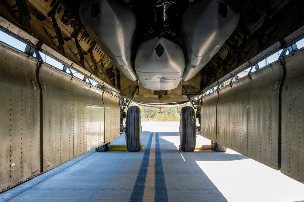 The image shows a view from directly beneath a large aircraft, looking along the underside fuselage toward the main landing gear. The perspective reveals the aircraft's belly structure, including riveted aluminum skin panels, structural framing, and what appears to be a weapons bay or equipment pod mounted to the underside. The aircraft is parked on a concrete surface, likely at an airfield or museum, with vegetation visible in the background.