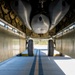The image shows a view from directly beneath a large aircraft, looking along the underside fuselage toward the main landing gear. The perspective reveals the aircraft's belly structure, including riveted aluminum skin panels, structural framing, and what appears to be a weapons bay or equipment pod mounted to the underside. The aircraft is parked on a concrete surface, likely at an airfield or museum, with vegetation visible in the background.