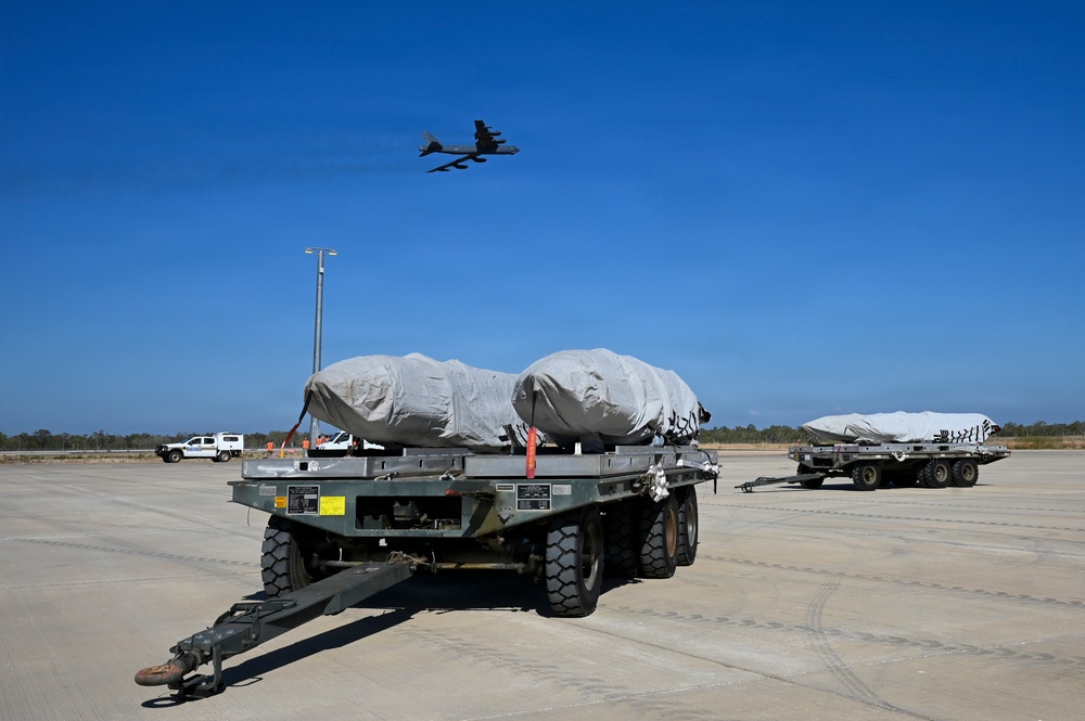 A military airfield scene showing two flatbed trailers loaded with large tarp-covered cargo secured with orange straps, positioned on a concrete apron. A large military aircraft, consistent with a B-52 Stratofortress, is visible flying overhead against a clear blue sky. A white utility vehicle and a light pole are visible in the background near a perimeter fence.